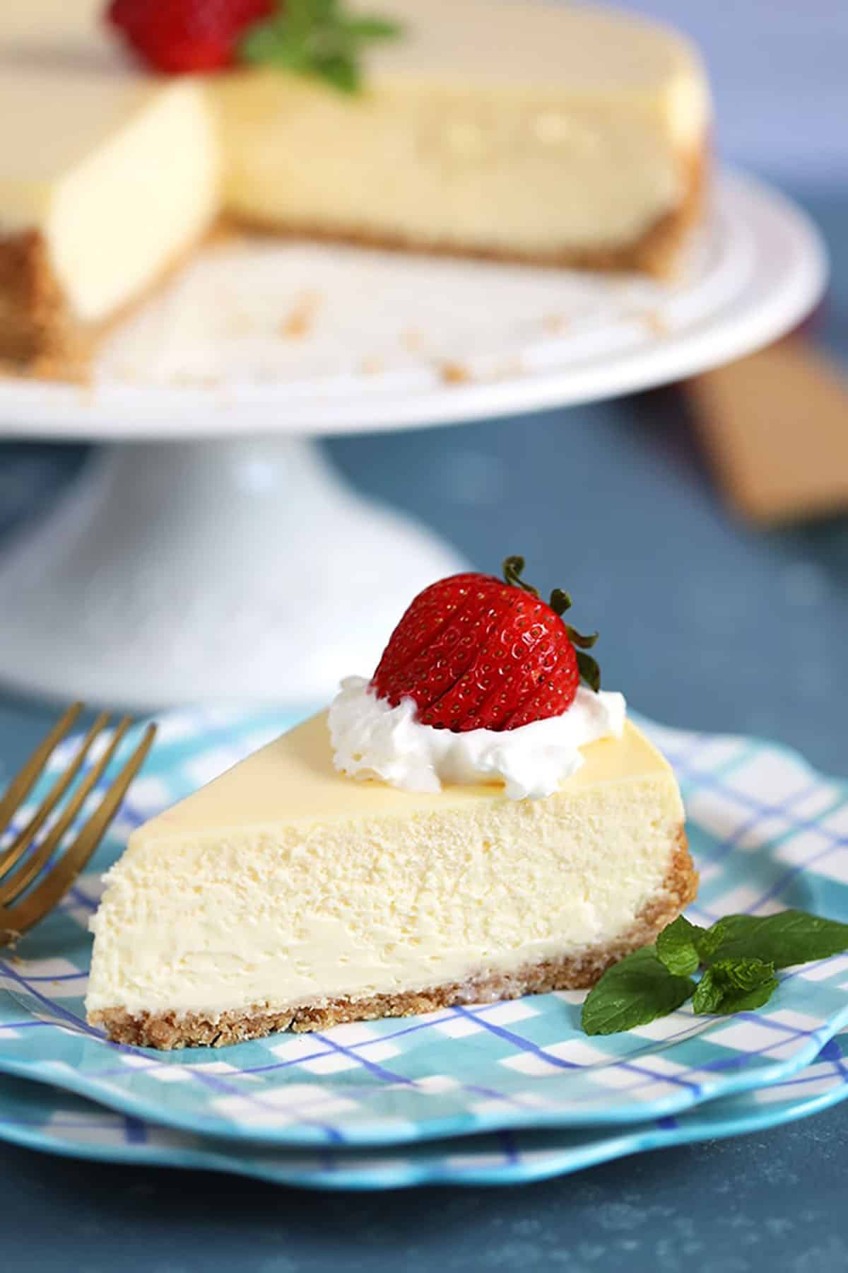 A slice of New York Cheesecake on a blue plaid plate with a strawberry and whipped cream on top and a sprig of mint.