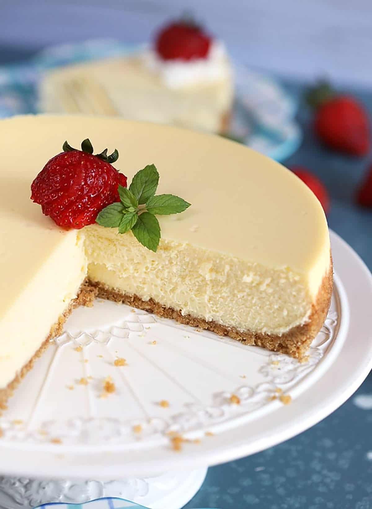 Close up of whole New York Cheesecake with a slice taken on a white cake plate.