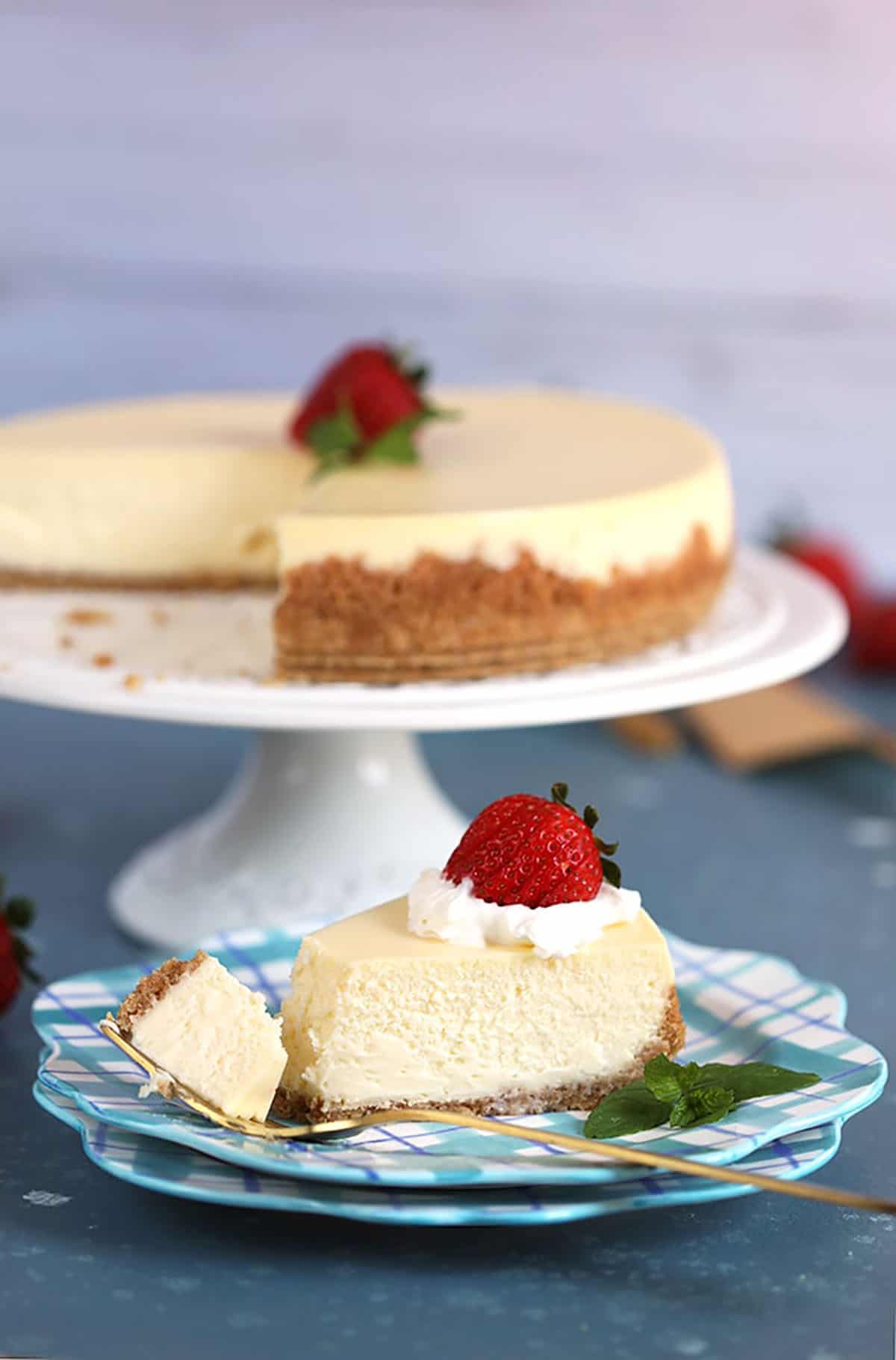 Shot of whole New York Cheesecake with a slice on a plaid plate and a fork with a bite taken.