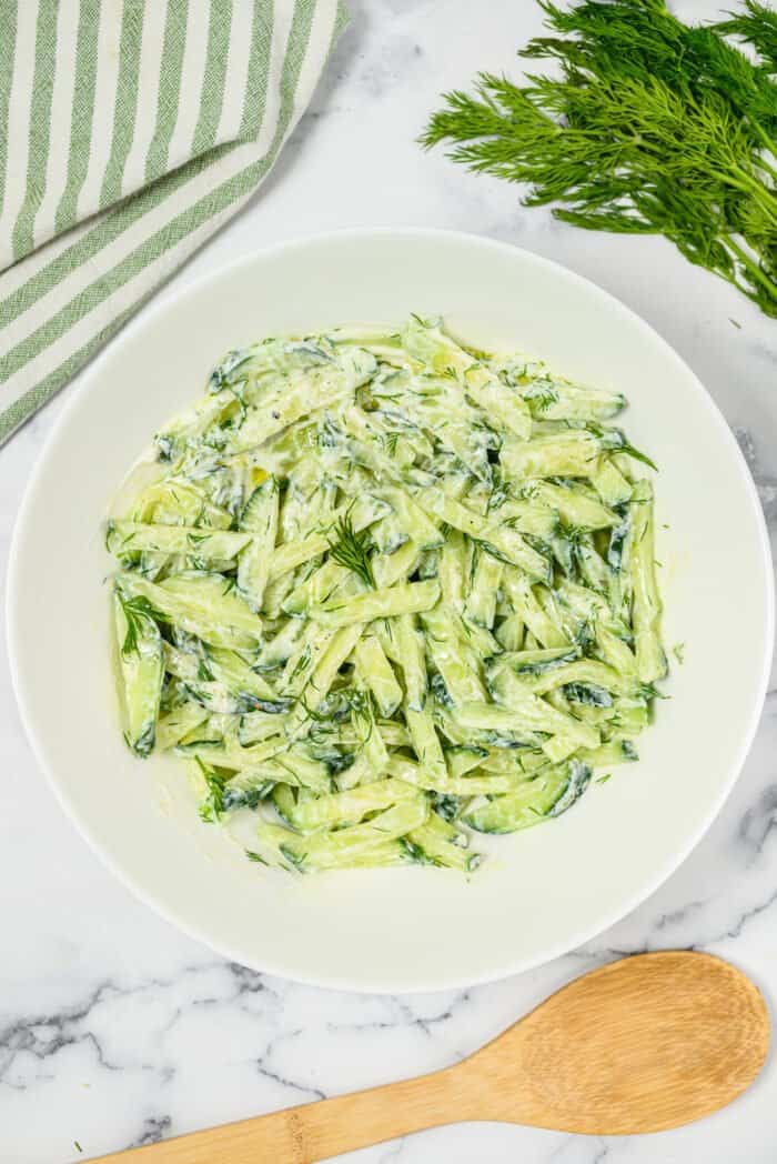 A serving of cucumber dill salad is presented on a white plate next to a wooden spoon.