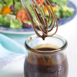 A whisk is held above a small jar of balsamic vinaigrette.