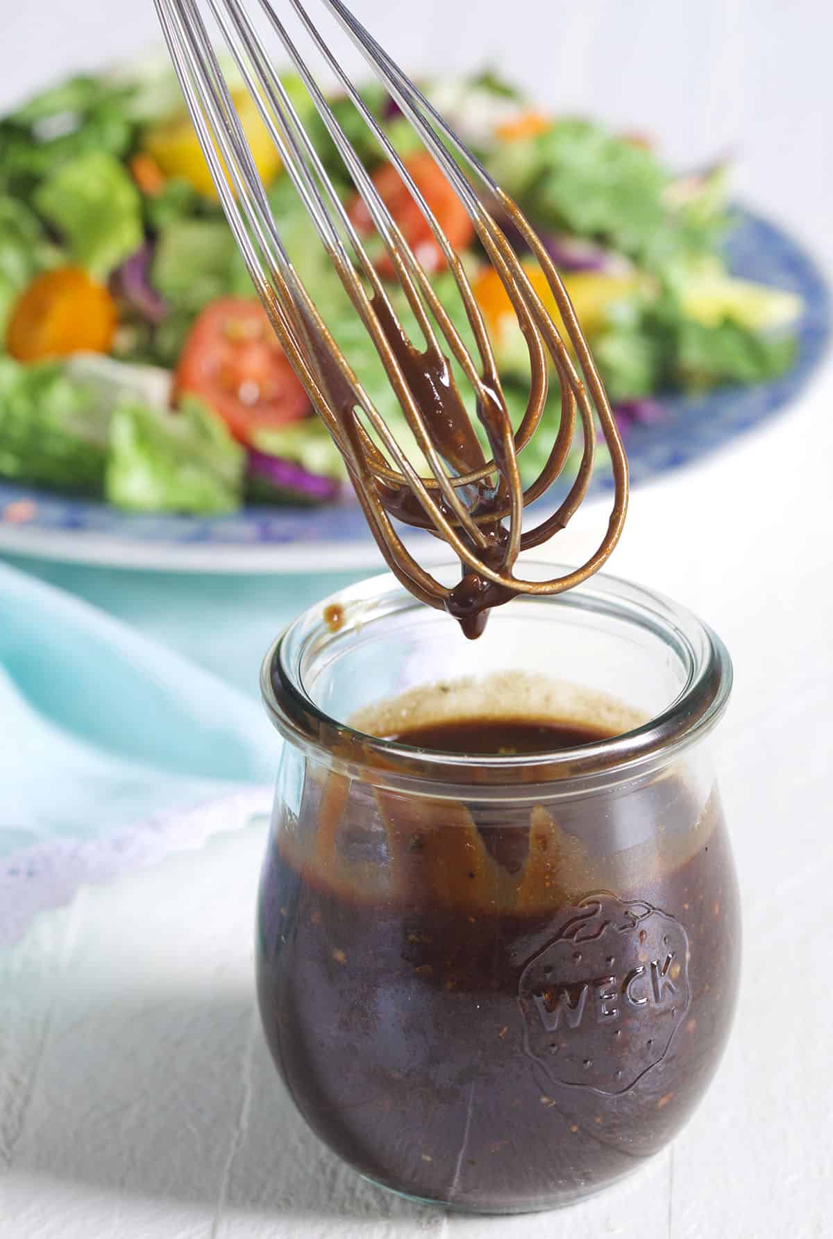 A whisk is held above a small jar of balsamic vinaigrette. 