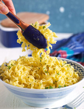 Buttered noodles in a blue and white bowl with tongs scooping out some noodles.