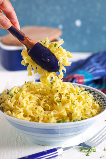 Buttered noodles in a blue and white bowl with tongs scooping out some noodles.