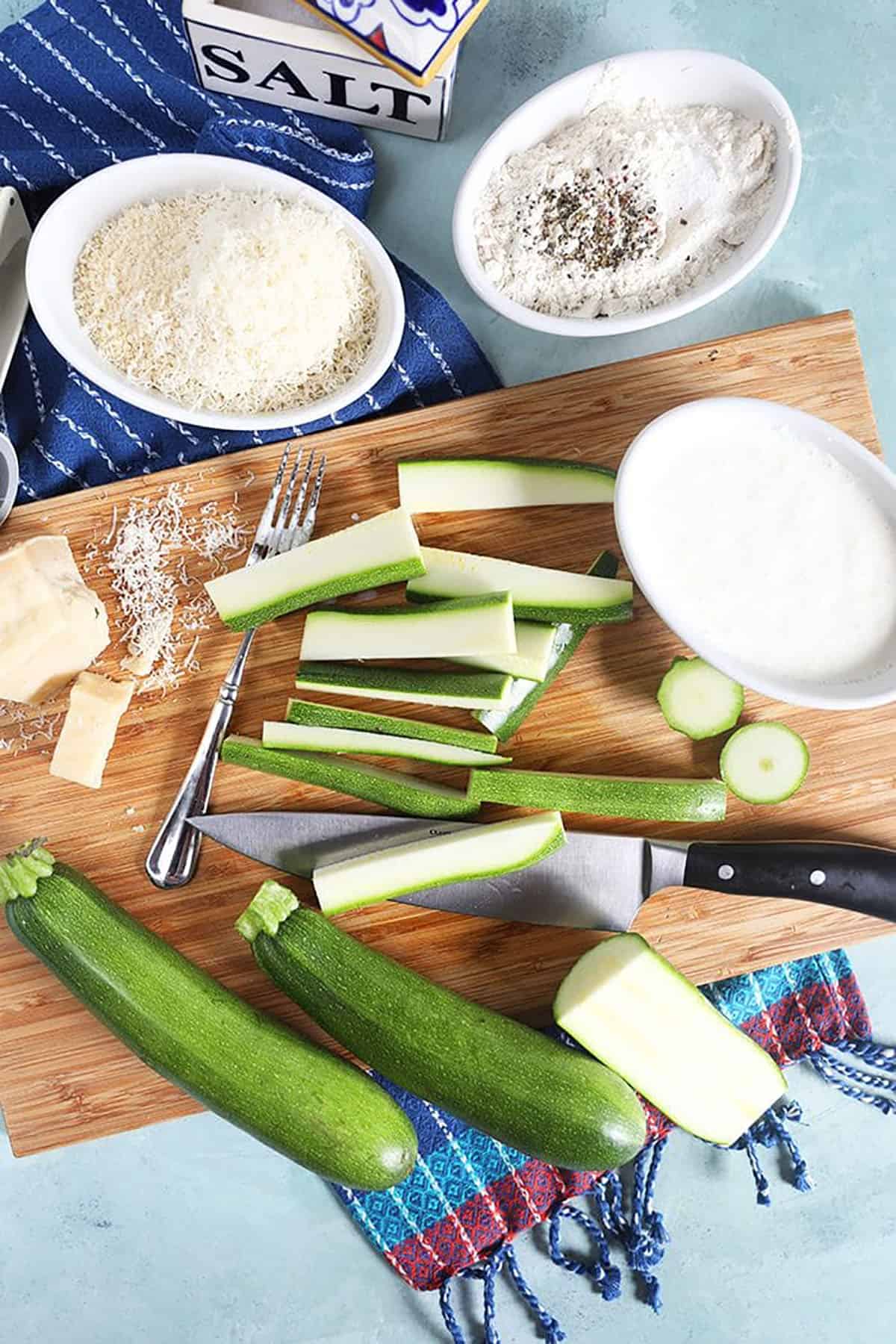 zucchini cut into sticks with three bowls with flour and breadcrumbs for dredging the zucchini.
