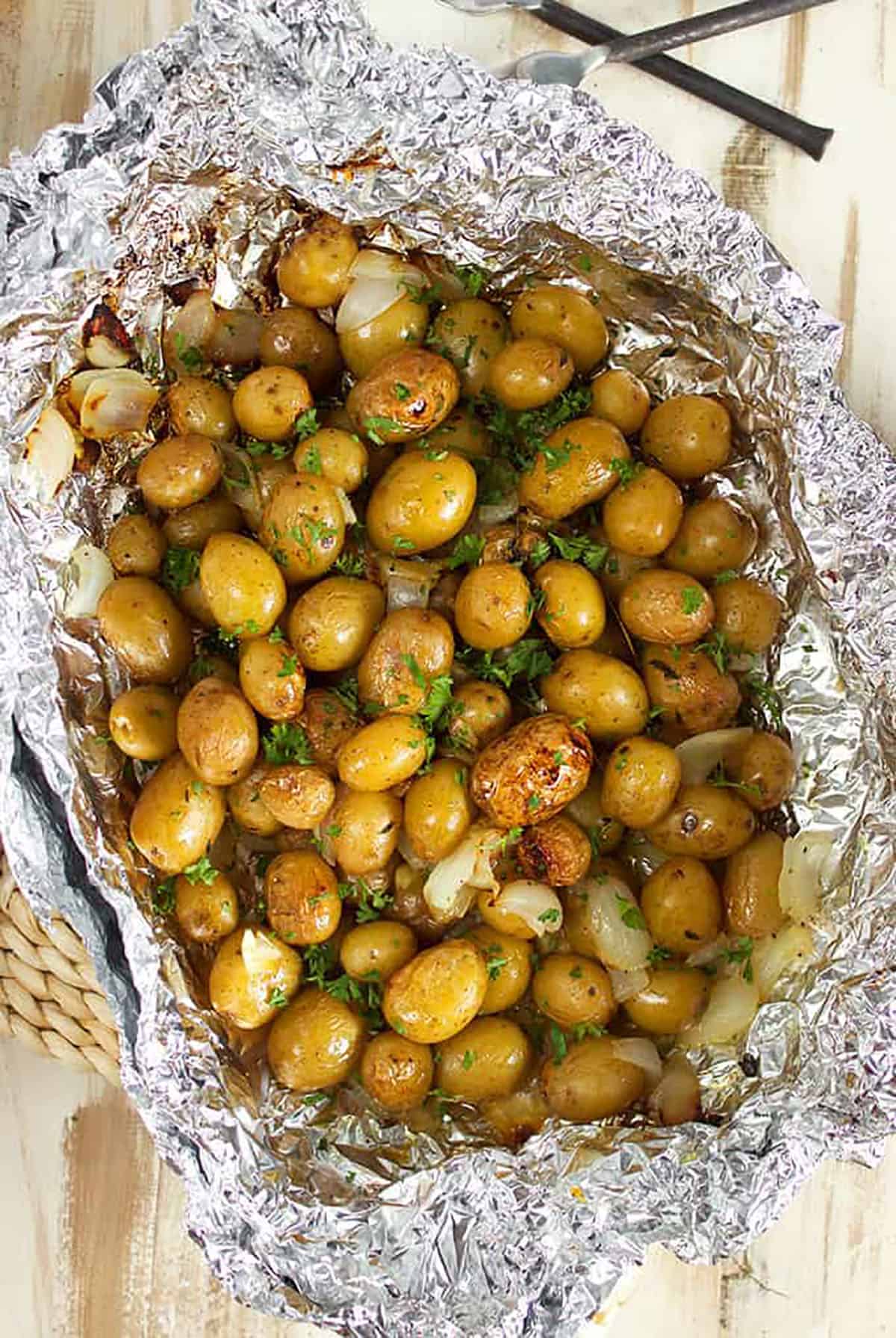 Grilled baby Yukon potatoes in a foil packet with garlic cloves on a wood background.