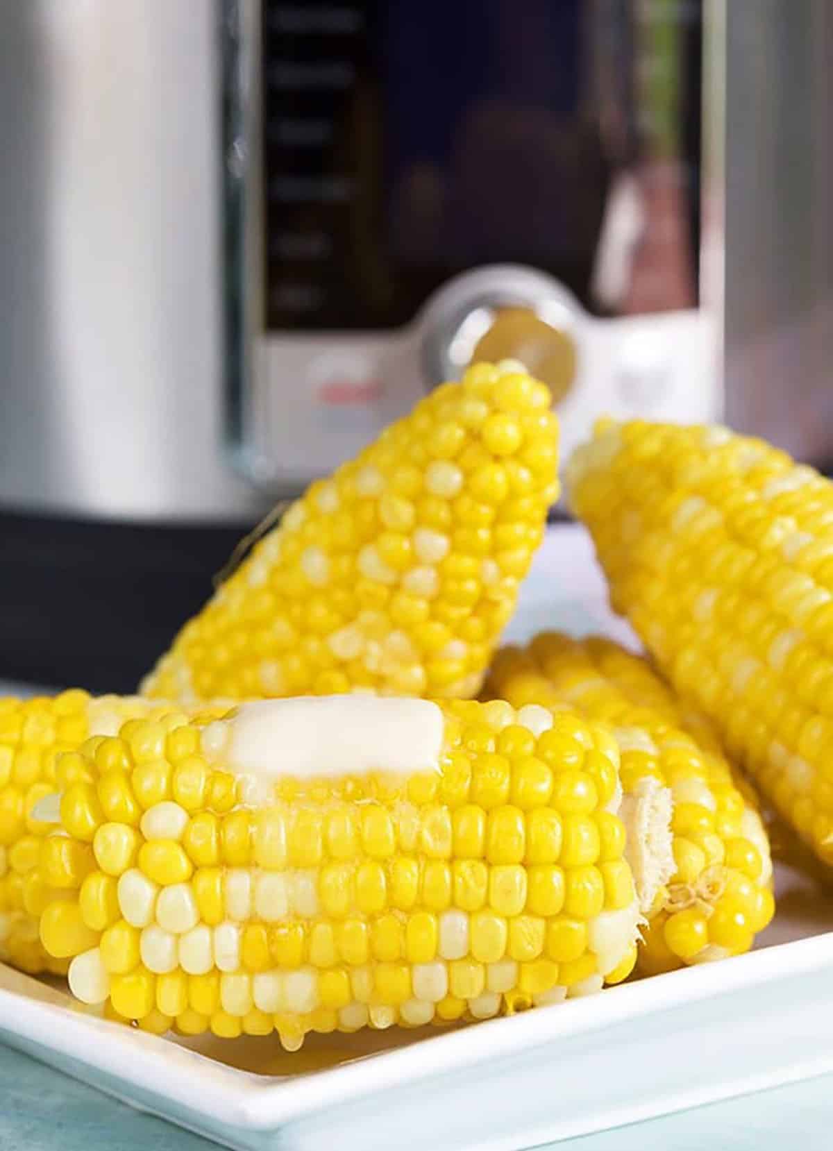 Platter of corn on the cob in front of Instant Pot with butter on top.