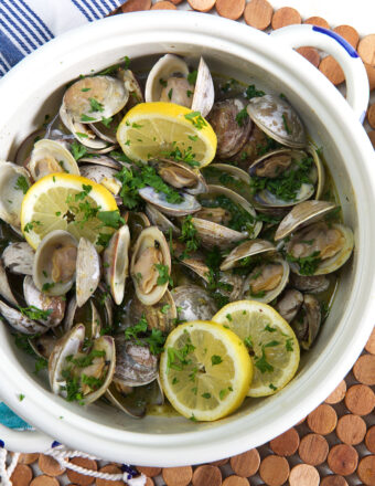 A white pot is filled with cooked clams, herbs, and lemon slices.