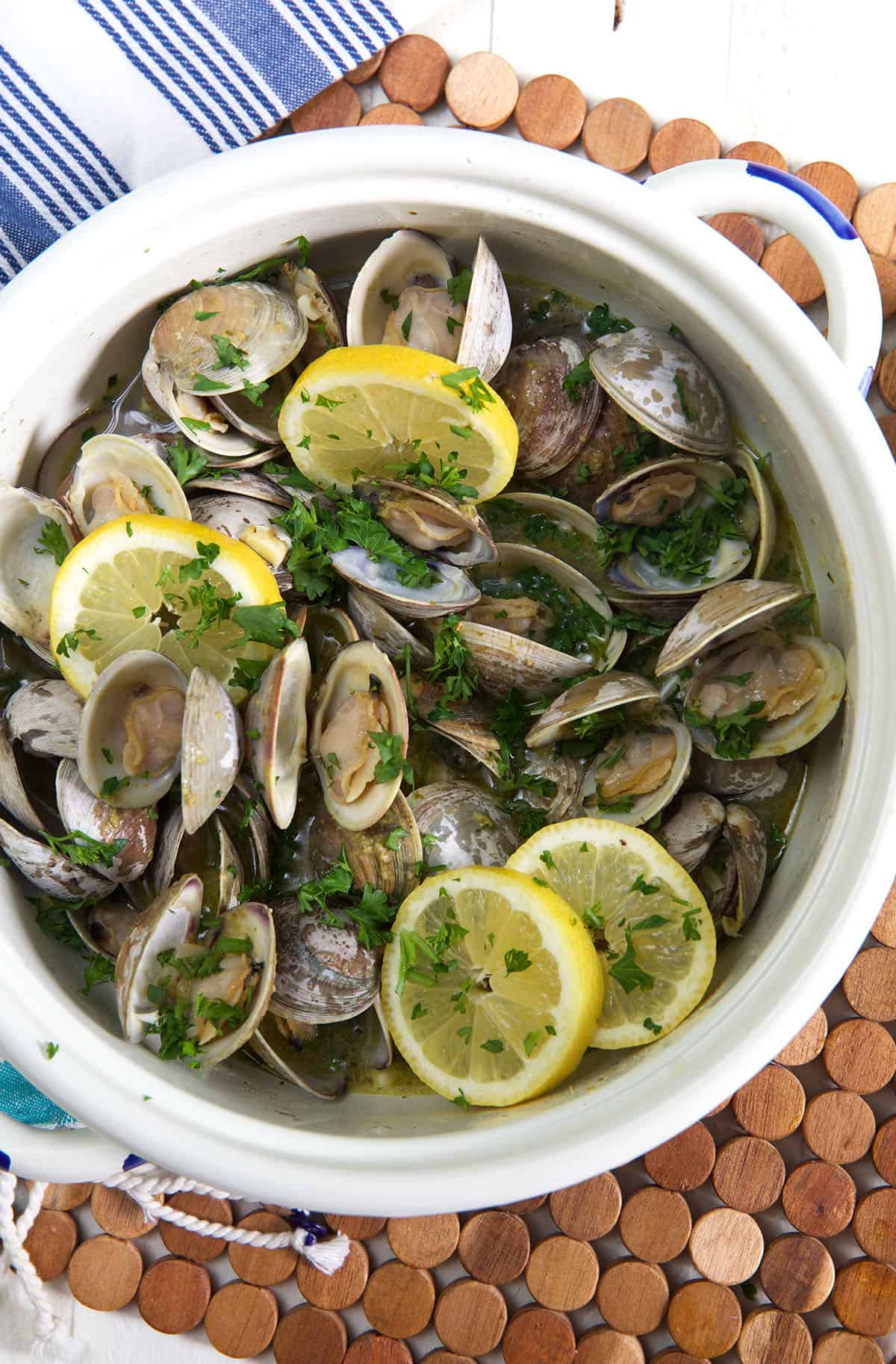 A white pot is filled with cooked clams, herbs, and lemon slices. 
