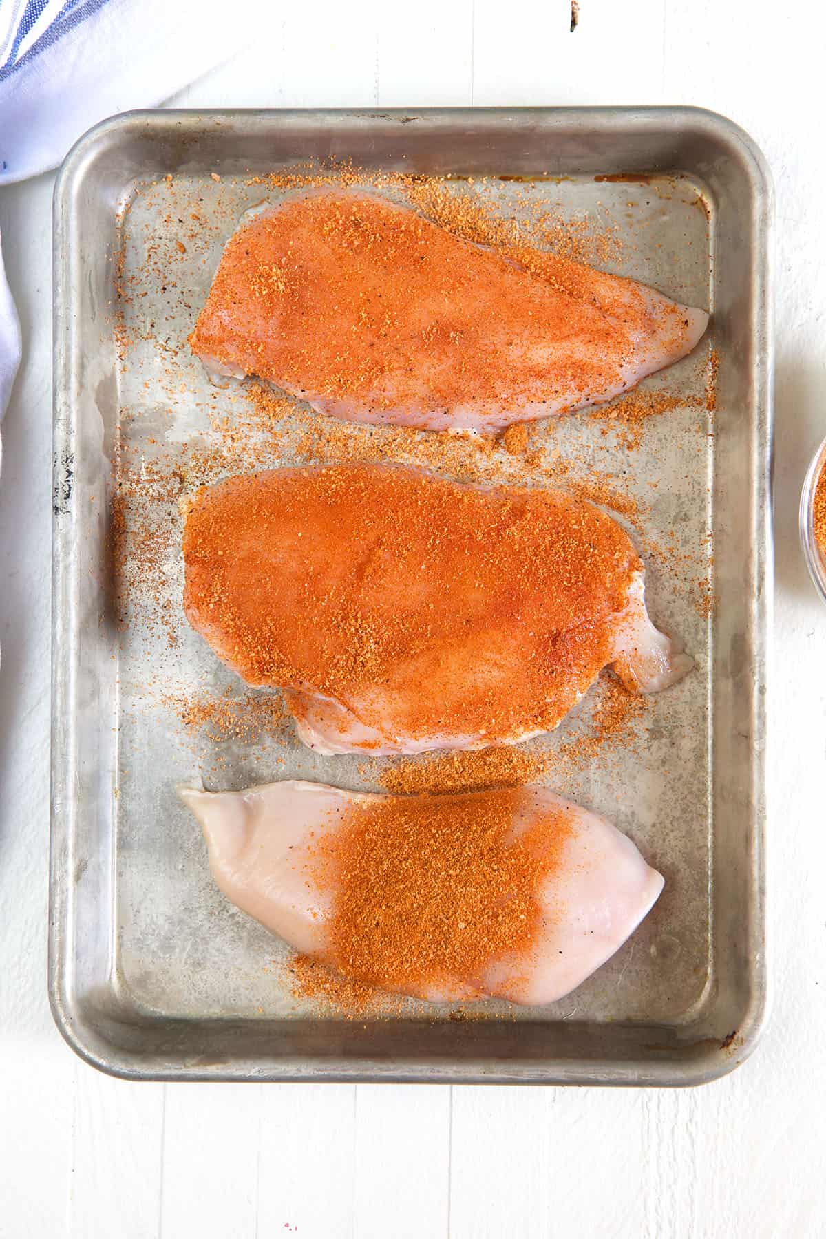 Three chicken breasts have been seasoned and are presented on a baking sheet.