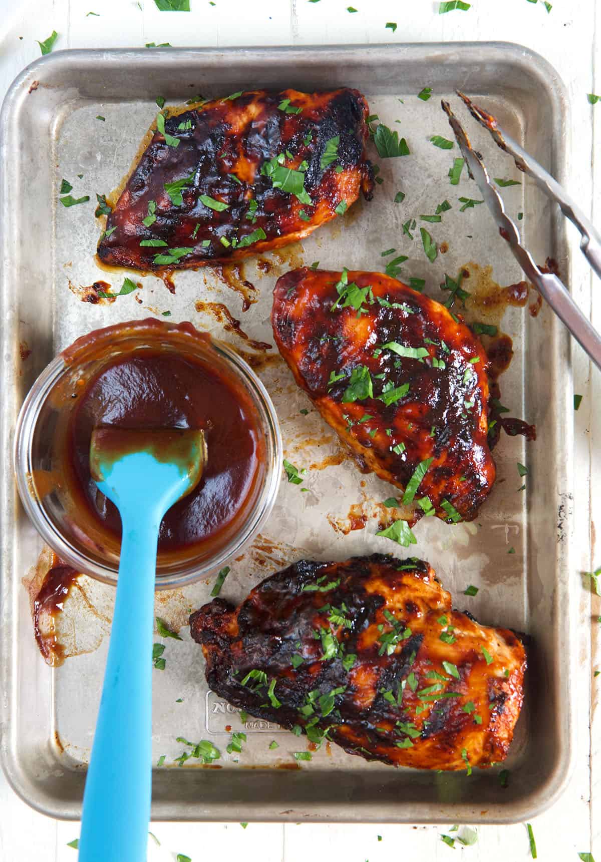 Three chicken breasts are placed next to a small bowl of extra BBQ sauce.