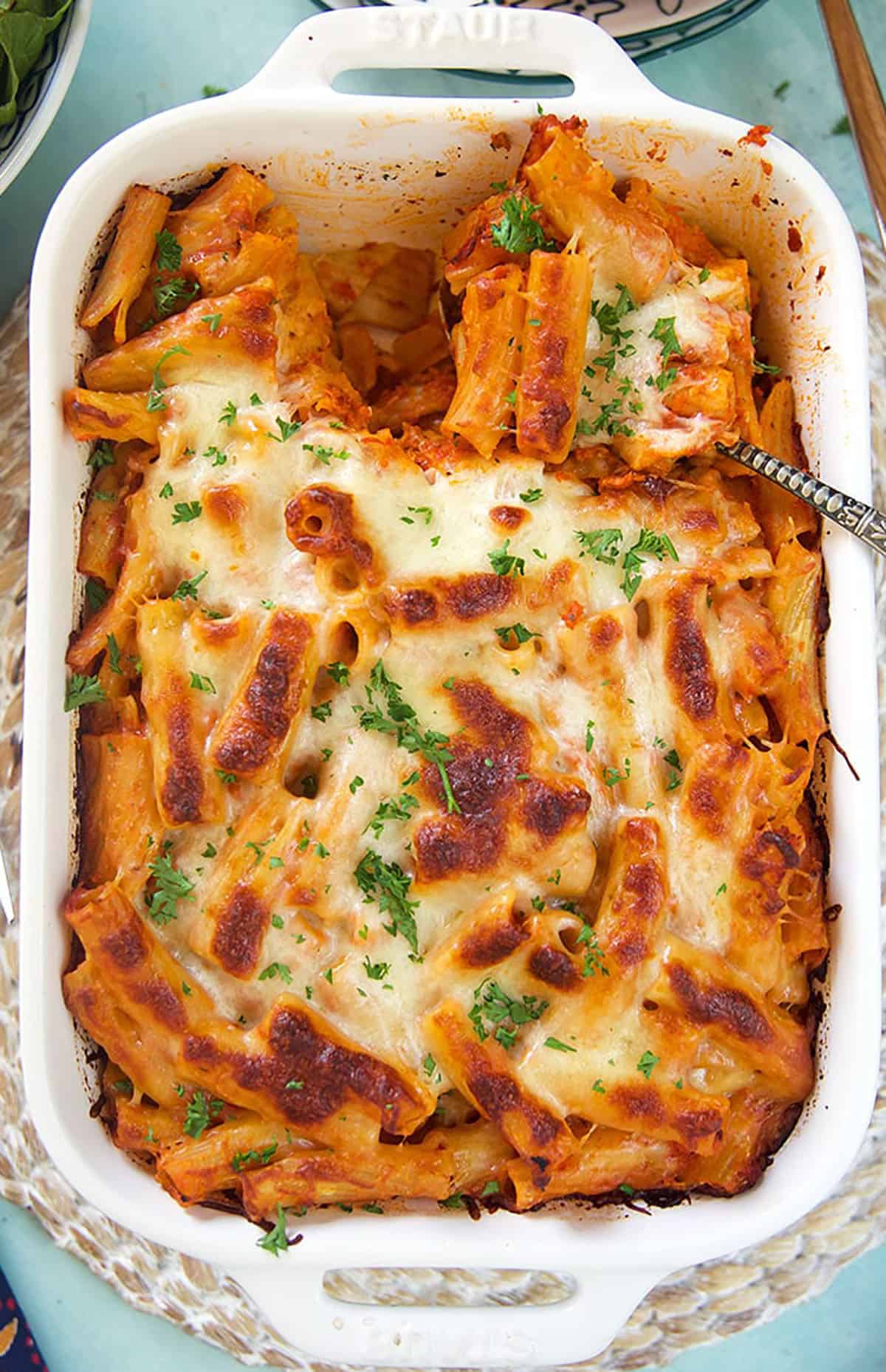 Overhead shot of baked rigatoni bolognese in a white baking dish on a blue background.