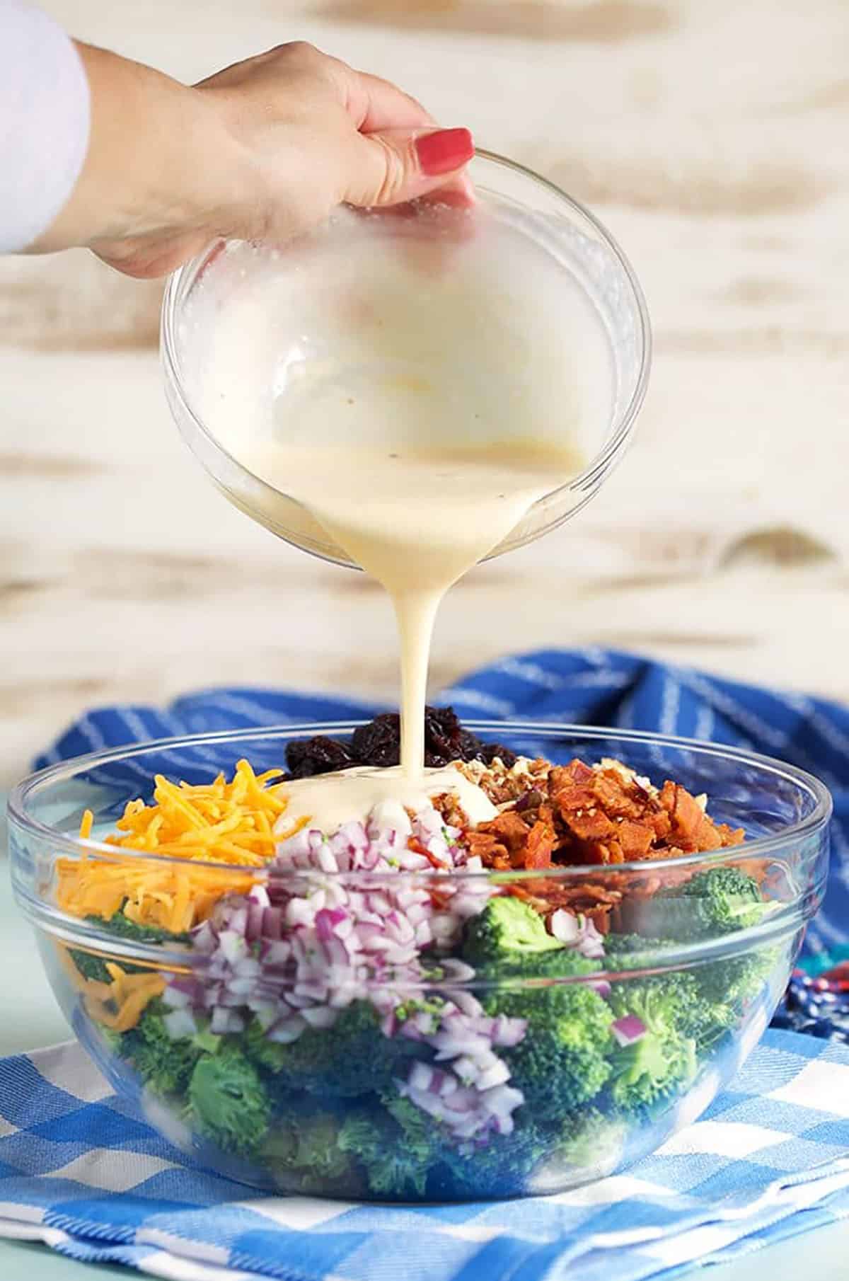 Broccoli salad dressing being poured over the salad in a glass bowl.