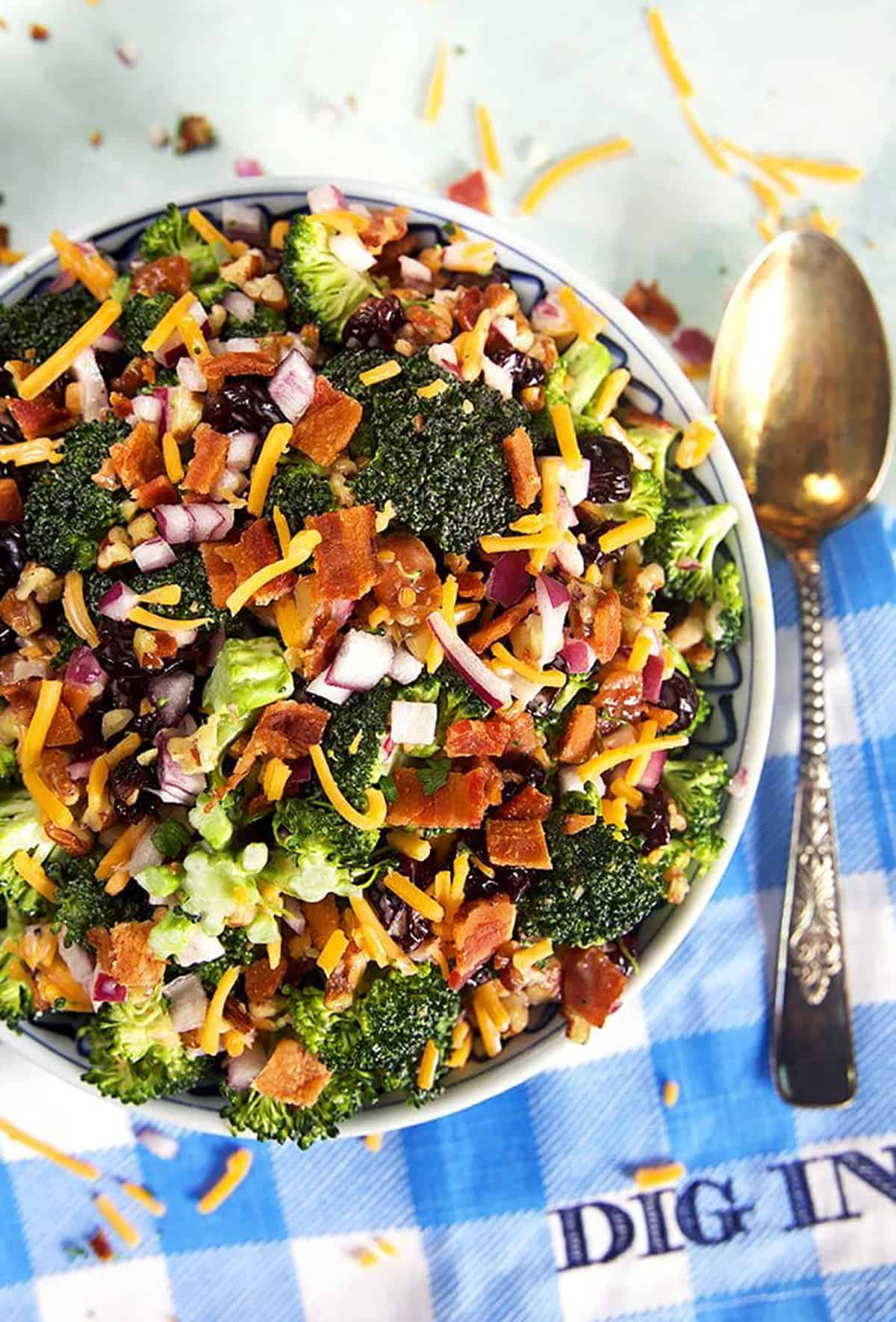 Overhead shot of broccoli salad with bacon on a blue and white checked napkin that says Dig In.