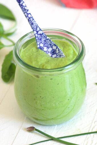 A small glass jar of green goddess dressing is placed on a white surface with a blue and white spoon.