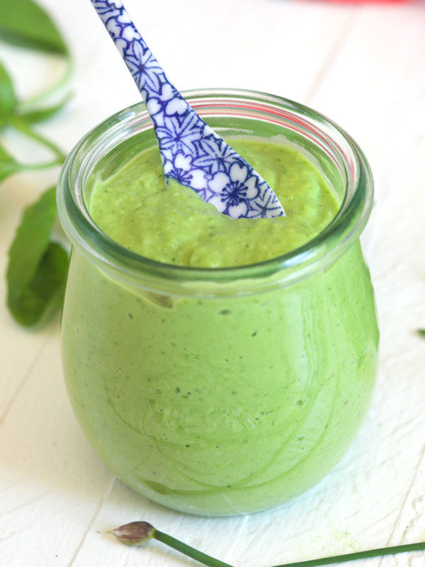 A small glass jar of green goddess dressing is placed on a white surface with a blue and white spoon.