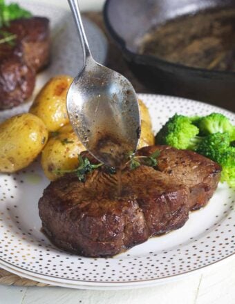 A spoon is adding more pan juices to the top of a cooked steak.