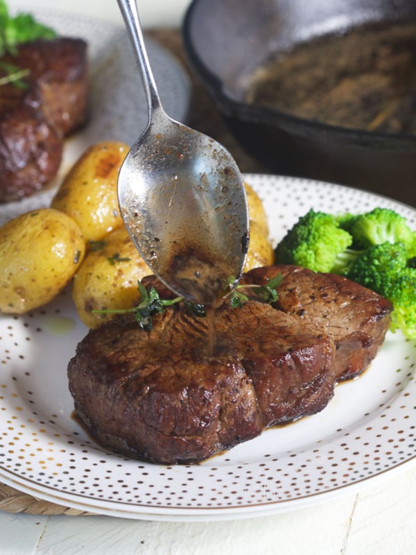 A spoon is adding more pan juices to the top of a cooked steak.