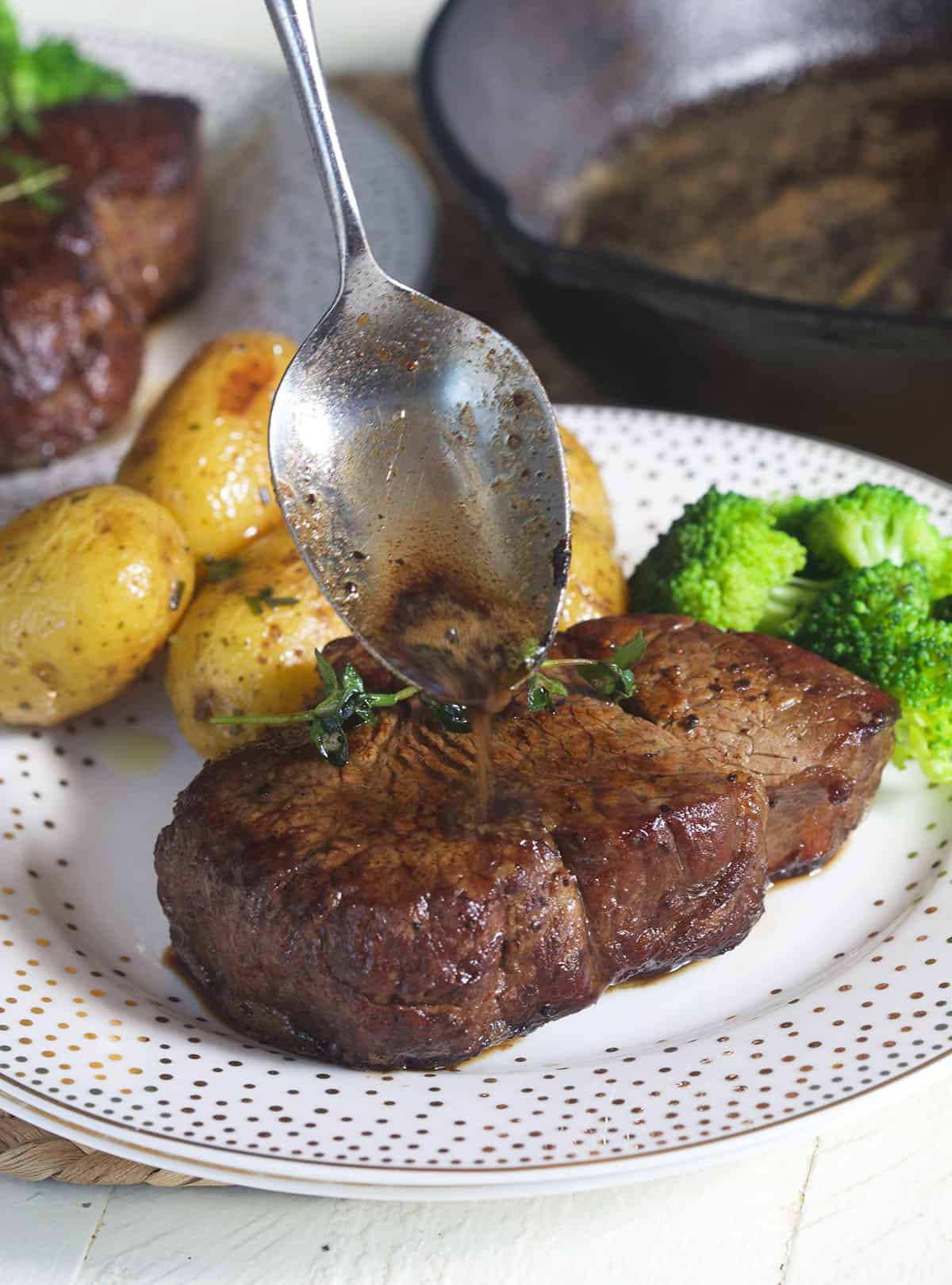 A spoon is adding more pan juices to the top of a cooked steak.