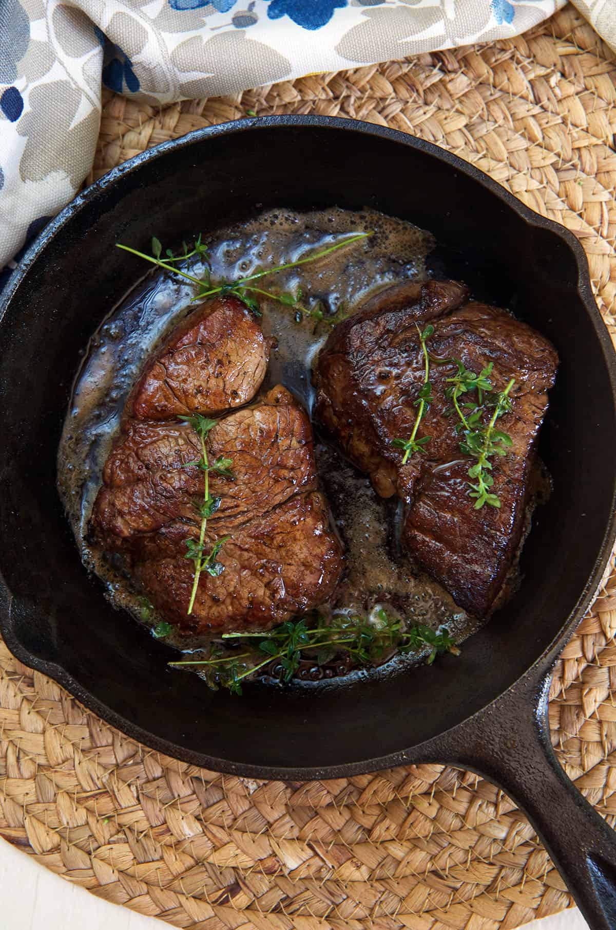 Two cooked steaks are presented with herbs in a black skillet.