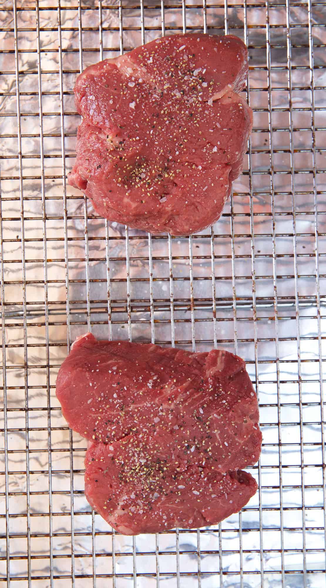 Two raw seasones steaks are placed on a wire rack.