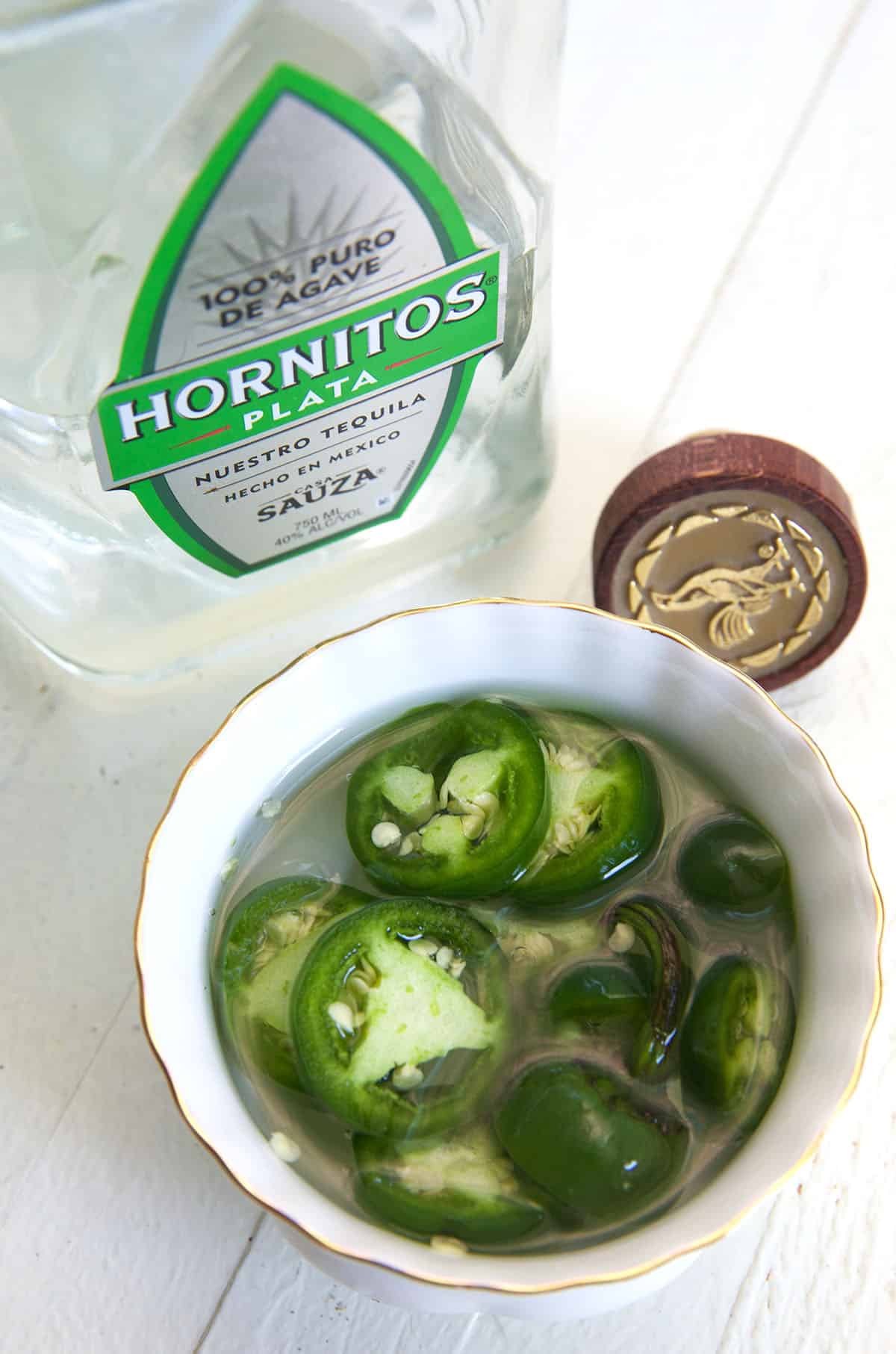 Jalapenos and tequila are placed next to each other on a white surface.