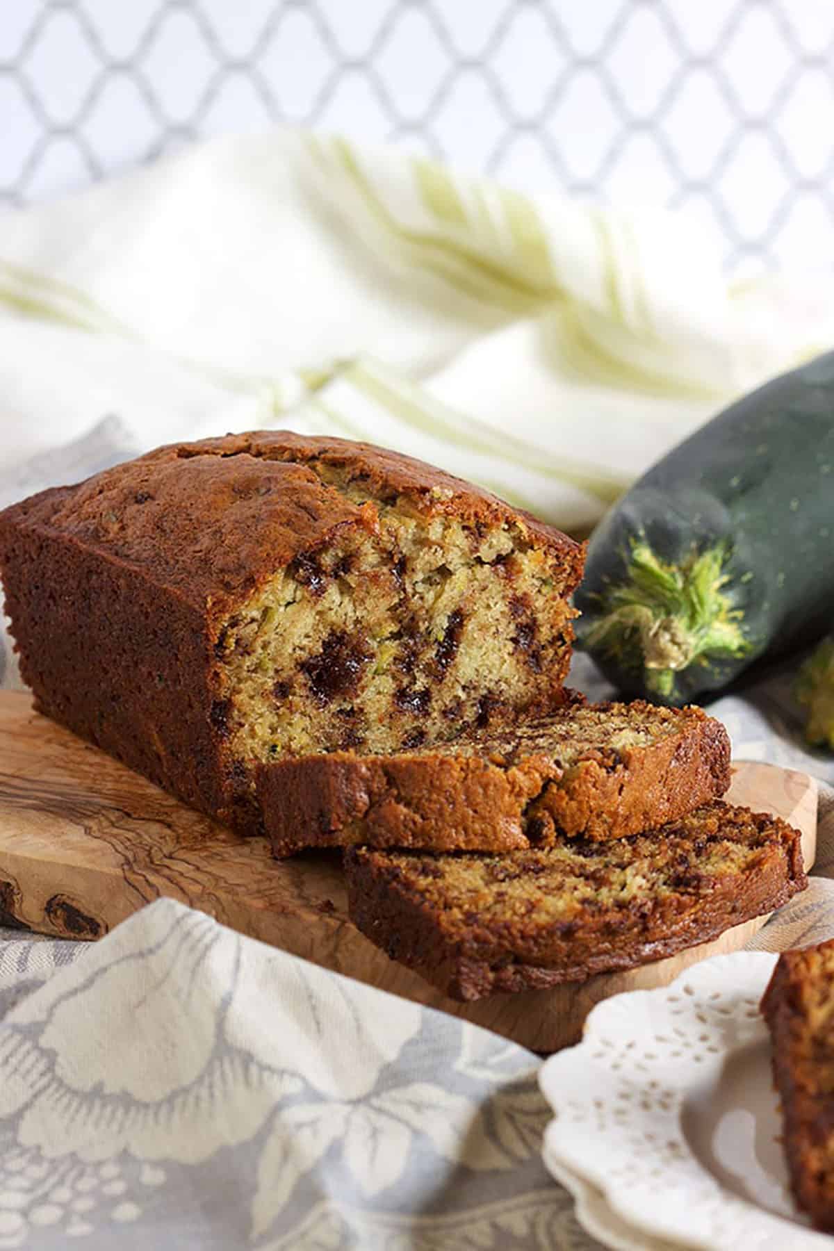 Zucchini Banana Bread with chocolate chips on an olive wood board with two slices in front of it and a raw zucchini in the background.