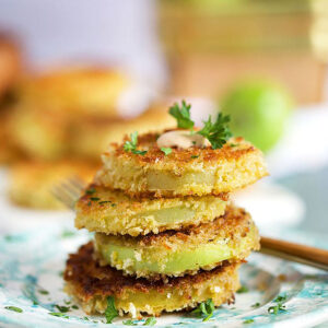 A stack of fried green tomatoes on plates with green tomatoes in the background in a basket