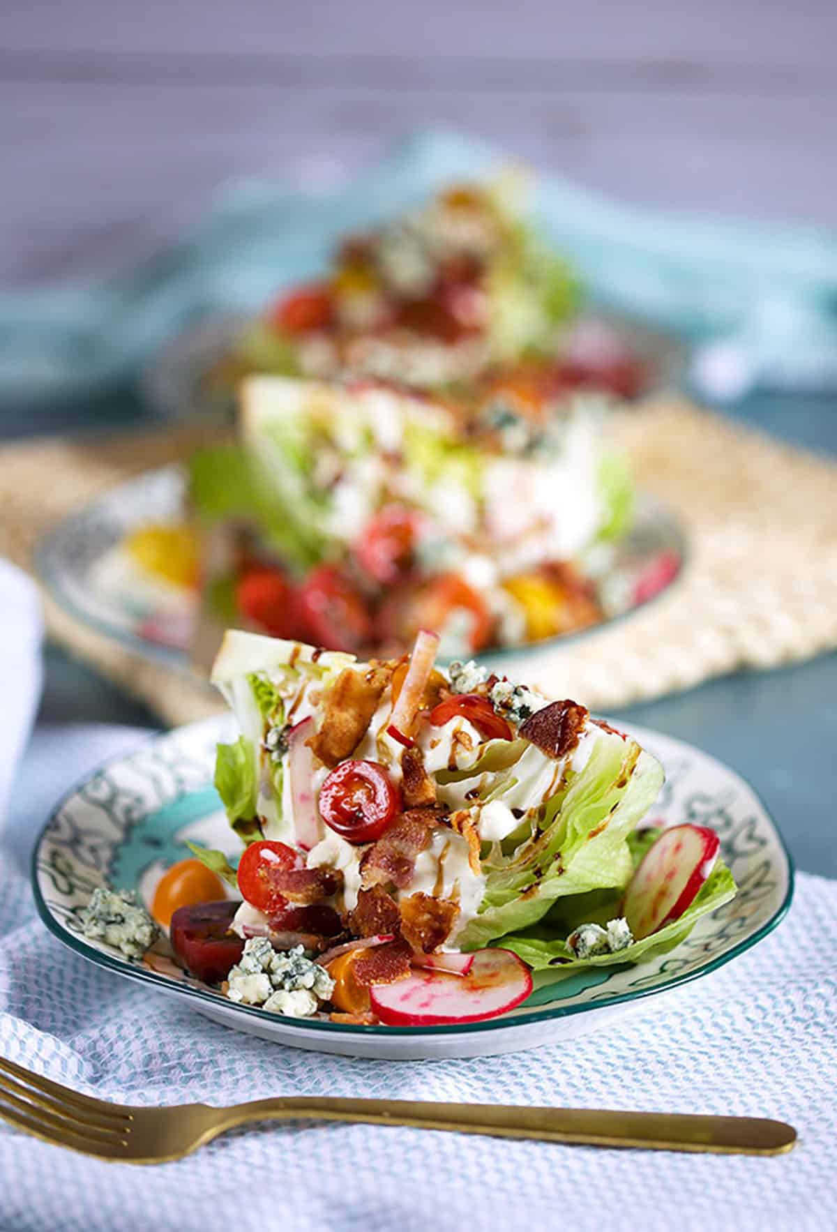 Row of three Loaded Iceberg Wedge Salads on white and blue plates with a gold fork on a blue background