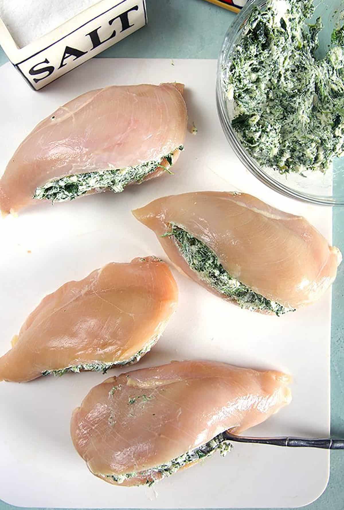 Overhead shot of four raw chicken breasts with spinach dip stuffing and a salt box on a white cutting board.