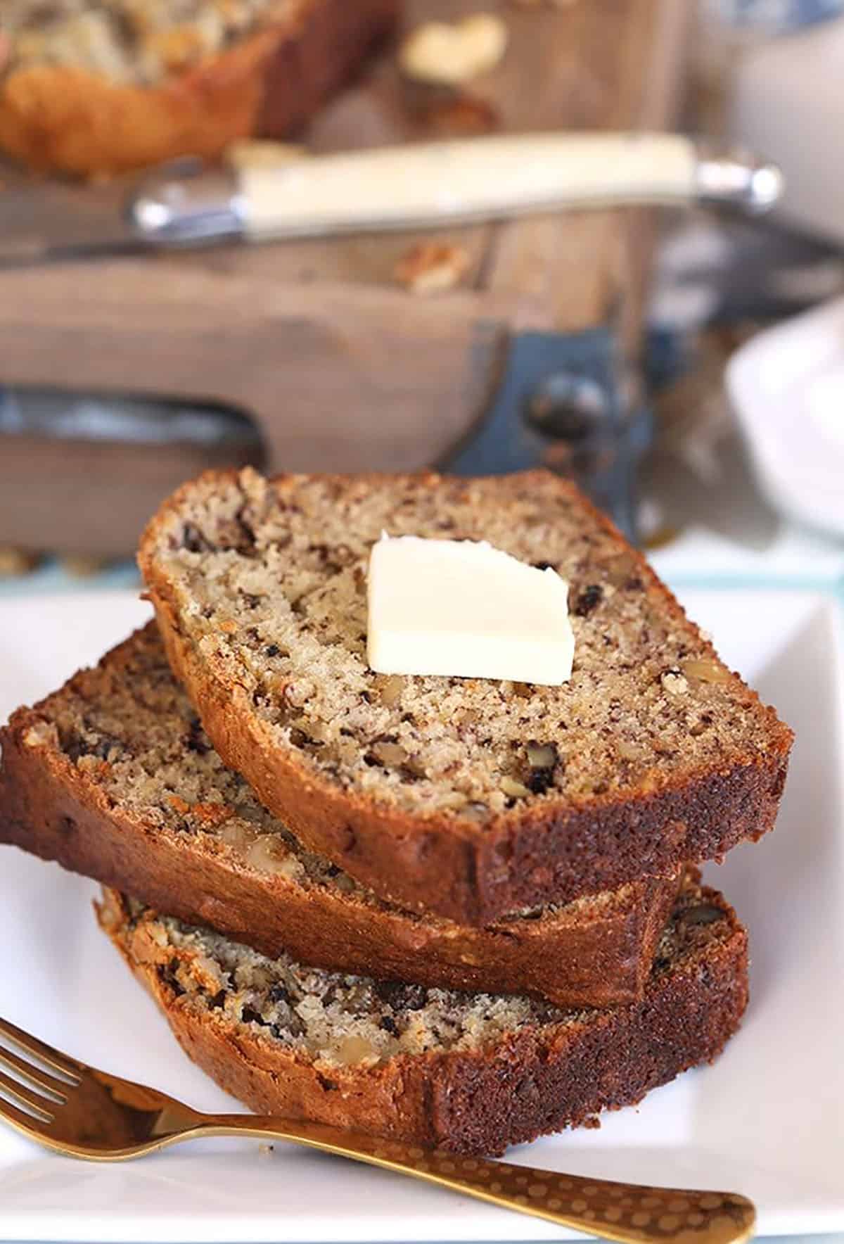 Three slices of banana nut bread on a white plate with a pat of butter on top and a gold fork.