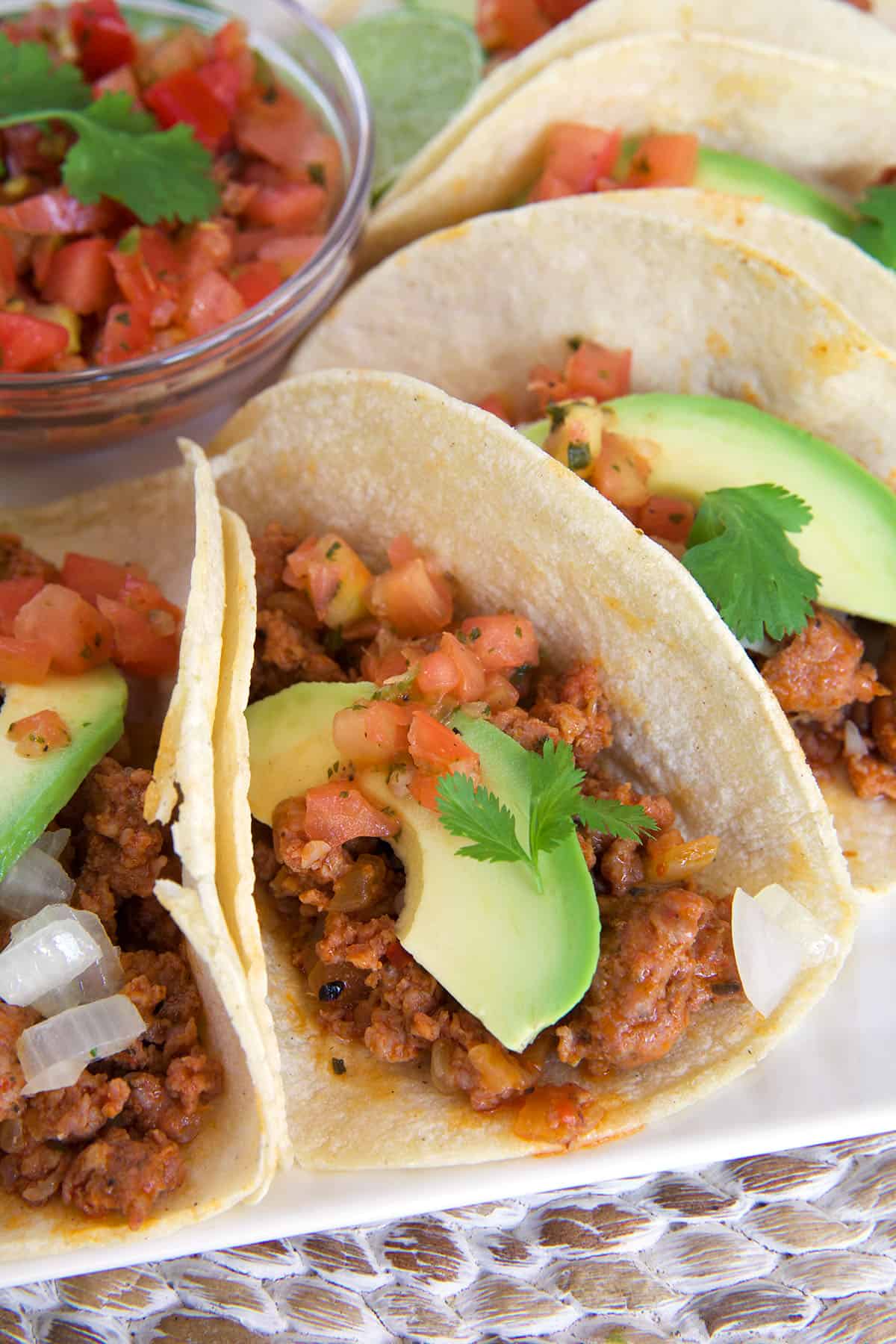 Several tacos are garnished and placed on a white plate with a small bowl filled with salsa.