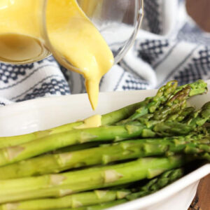 Hollandaise Sauce in a glass pitcher being poured over asparagus in a white dish.