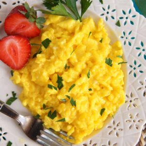 Scrambled eggs are placed on a white plate with sliced srawberries.