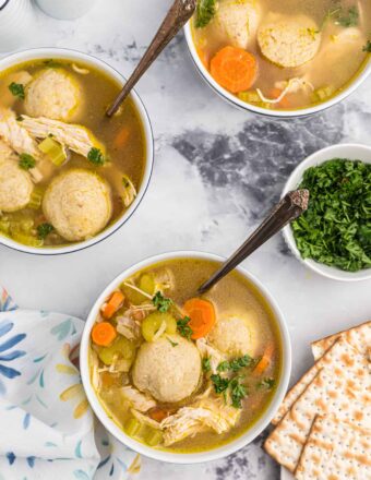 Three bowls of matzo ball soup are placed on a marble countertop.