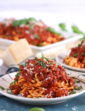 Pasta Bolognese on a white plate on a blue background with a wedge of parmesan in the background.