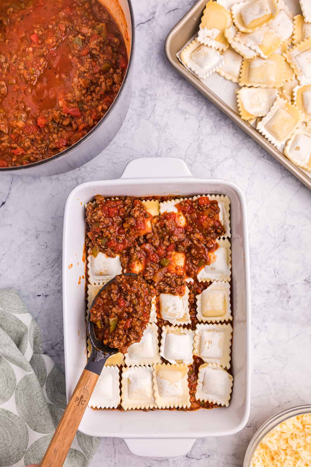 Meat sauce is being spread over a layer of raviolis in a white baking dish.