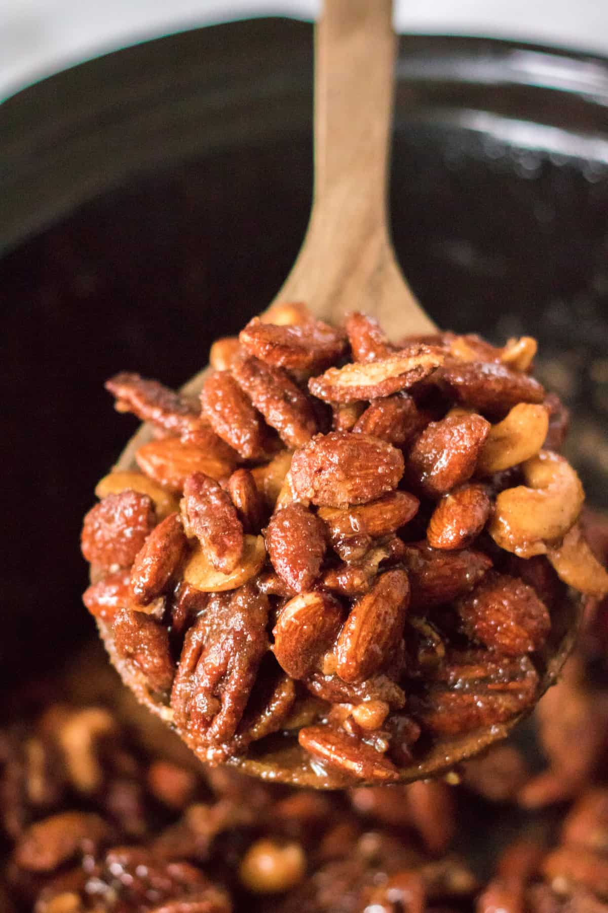 A wooden spoon is lifting a portion of cooked nuts from the bowl of a Crockpot.