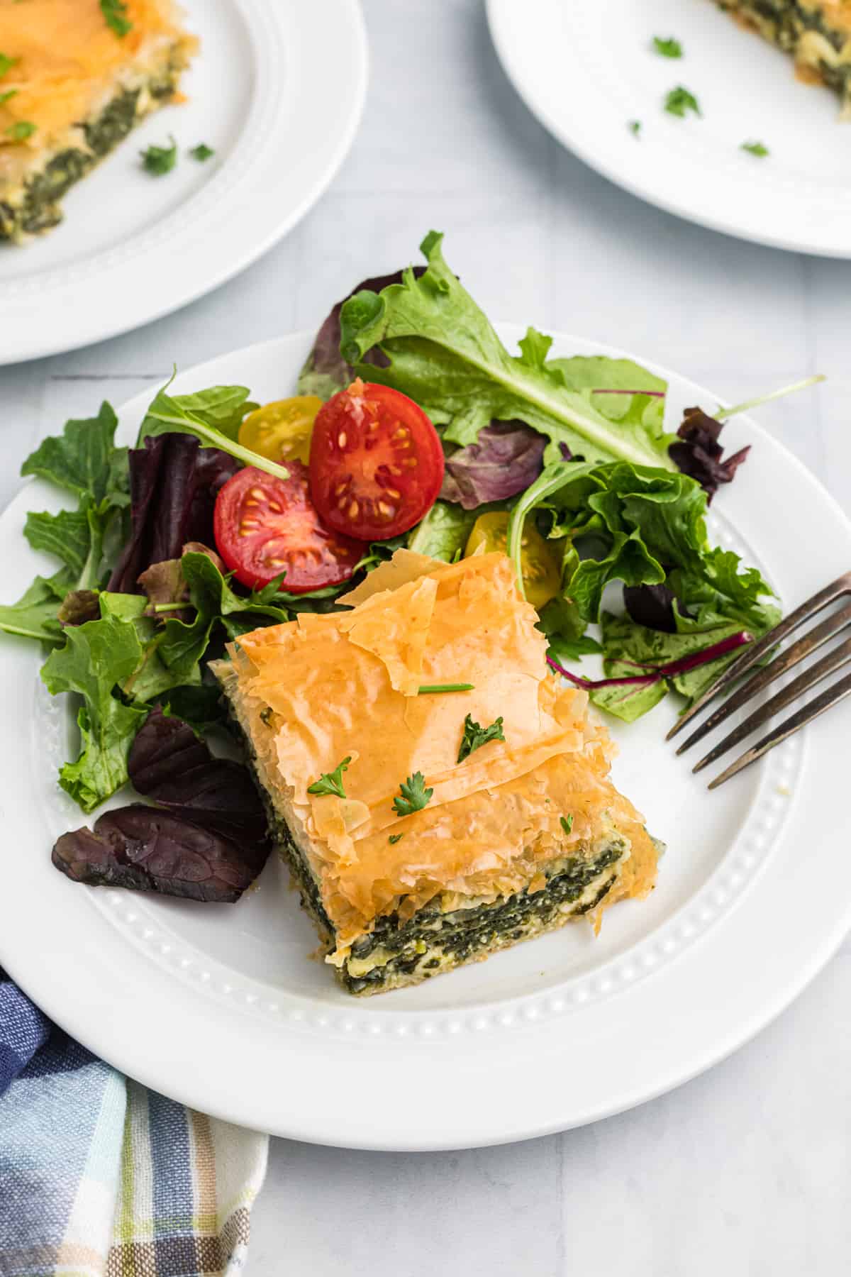 A piece of spanakopita is placed on a white plate next to salad.