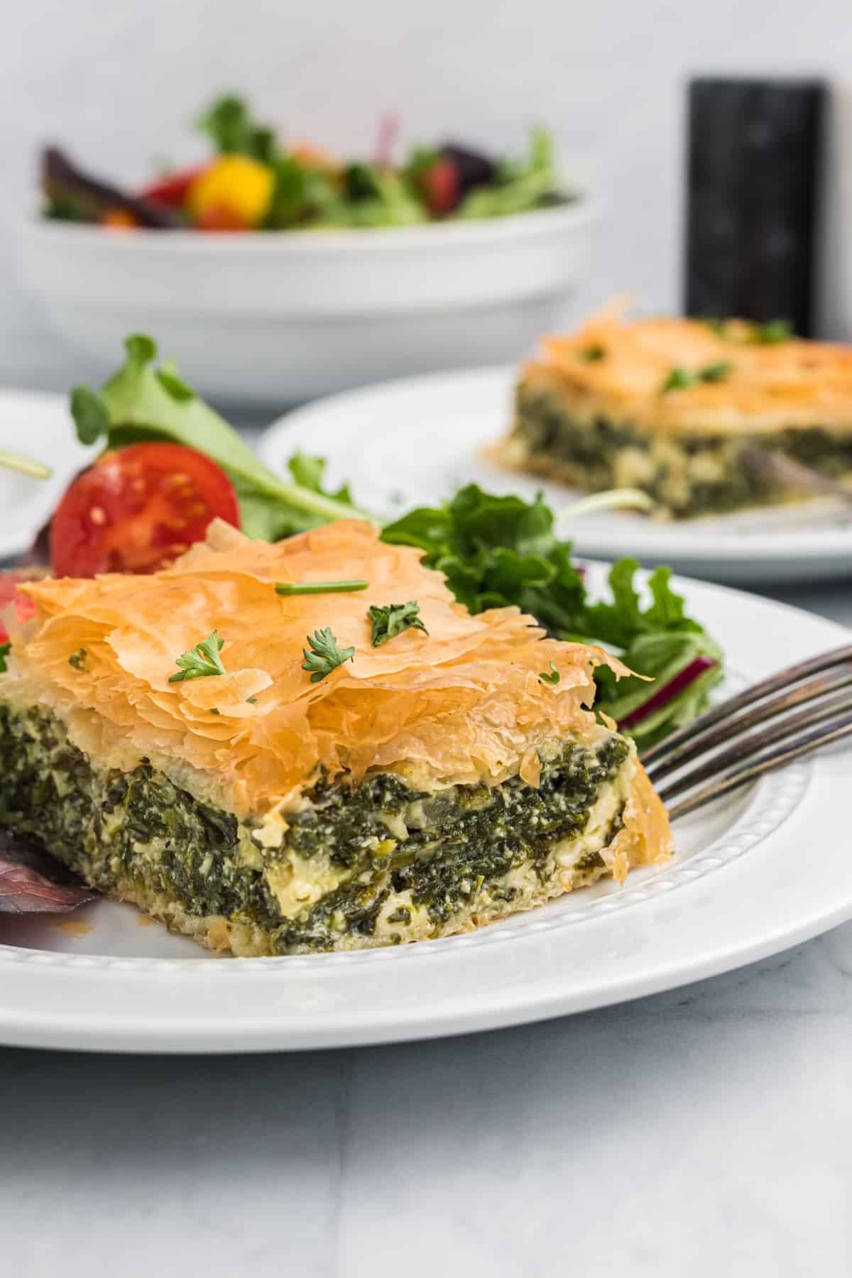 A slice of spanakopita is placed on a white plate next to a side salad.
