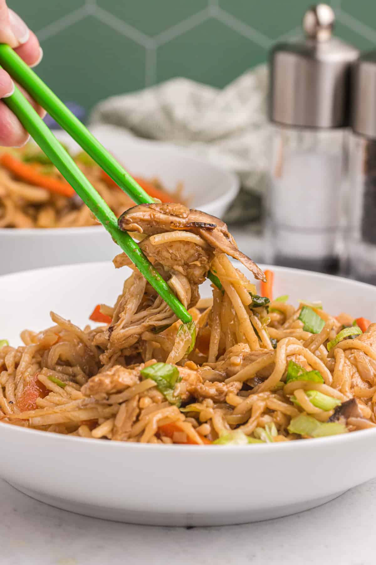 Yakisoba is being lifted from a white serving dish with green chopsticks.
