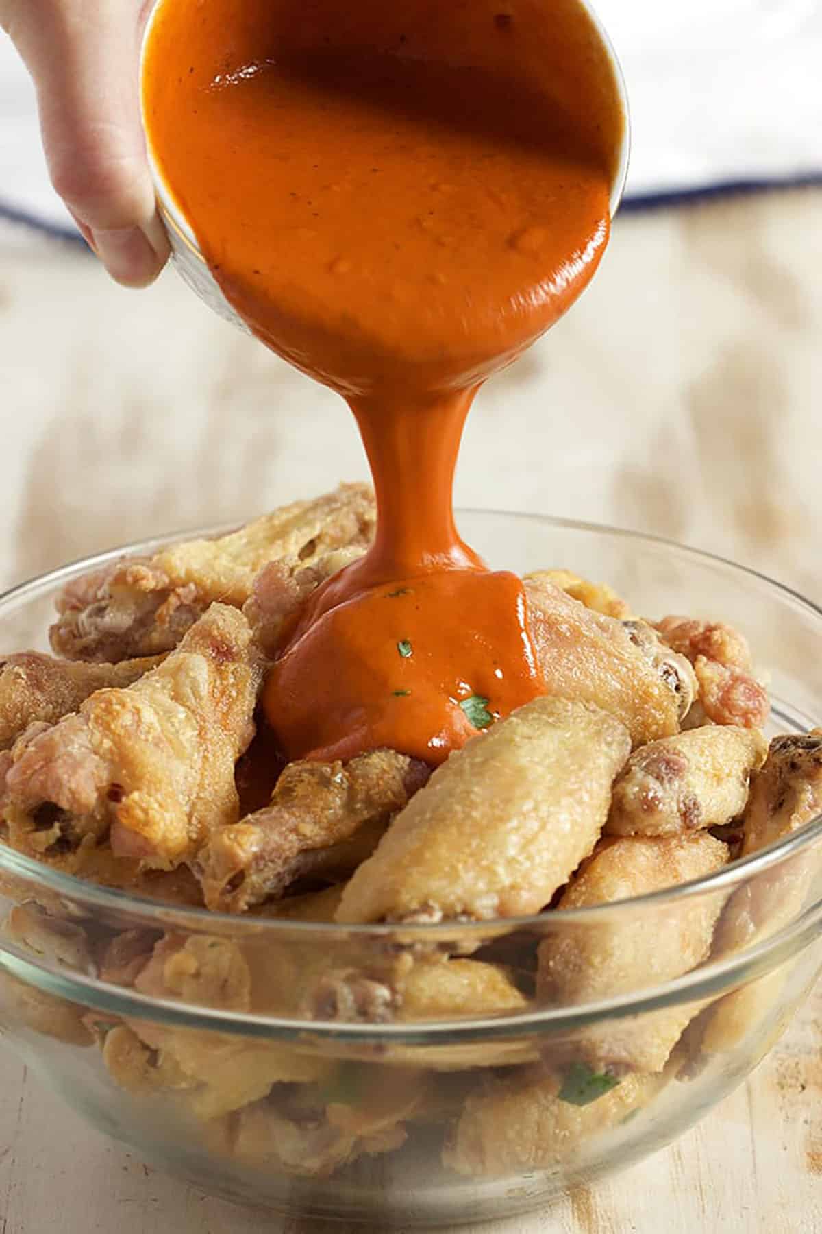crispy baked chicken wings in a bowl with buffalo sauce being poured over top.