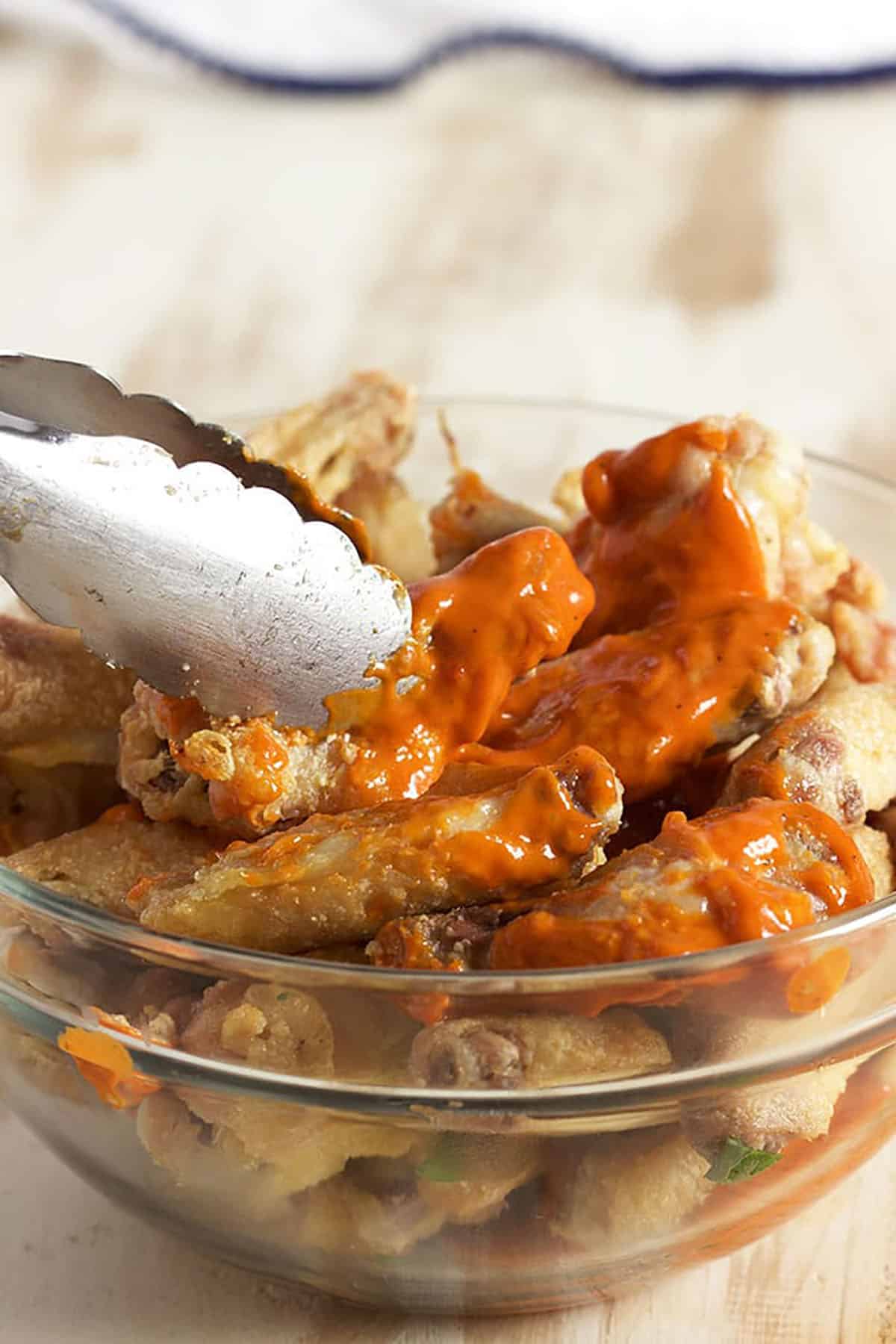 chicken wings being tossed with tongs in a bowl of buffalo sauce.