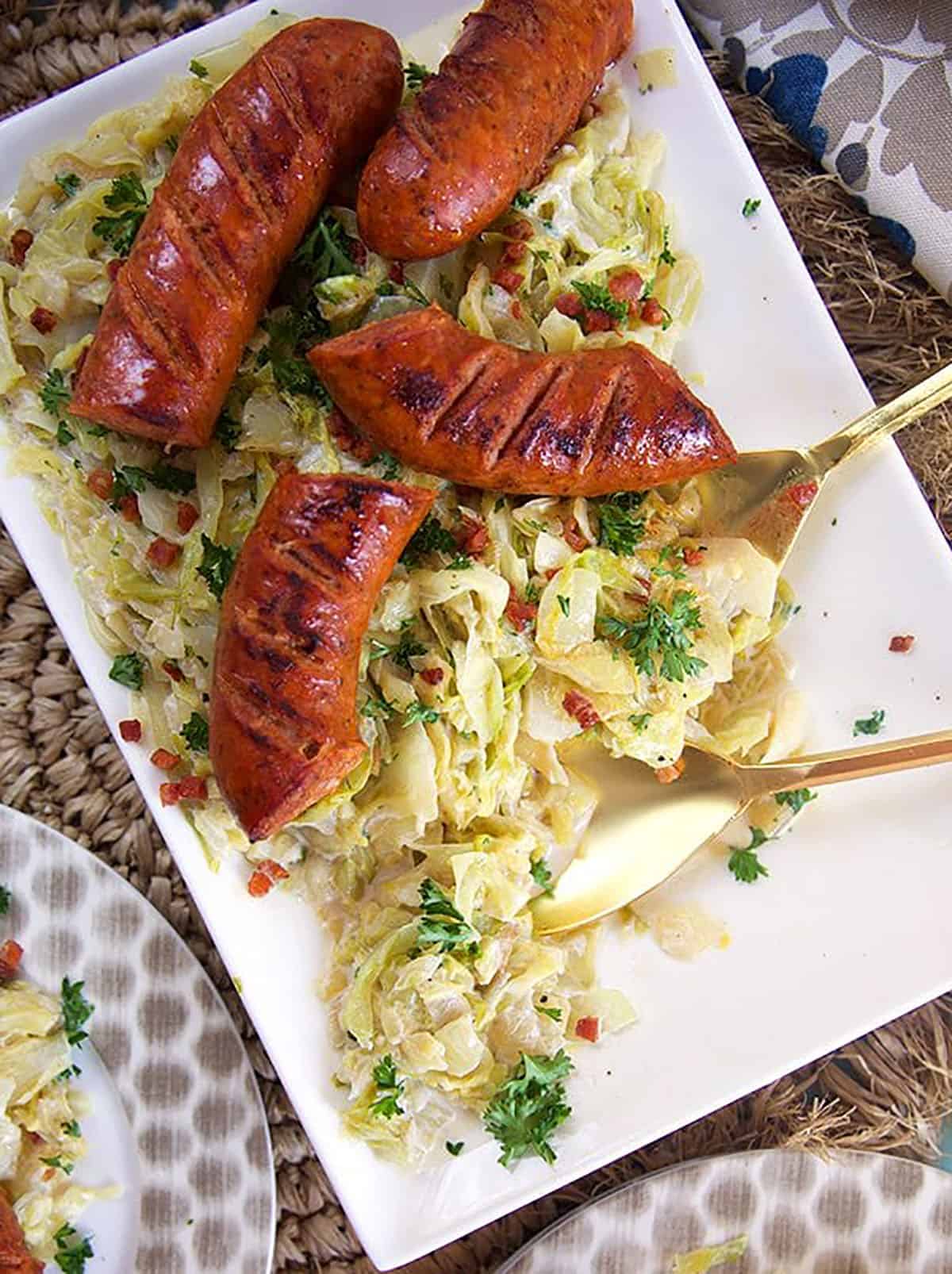 Overhead shot of fried cabbage and kielbasa on a white platter with a gold serving spoon.