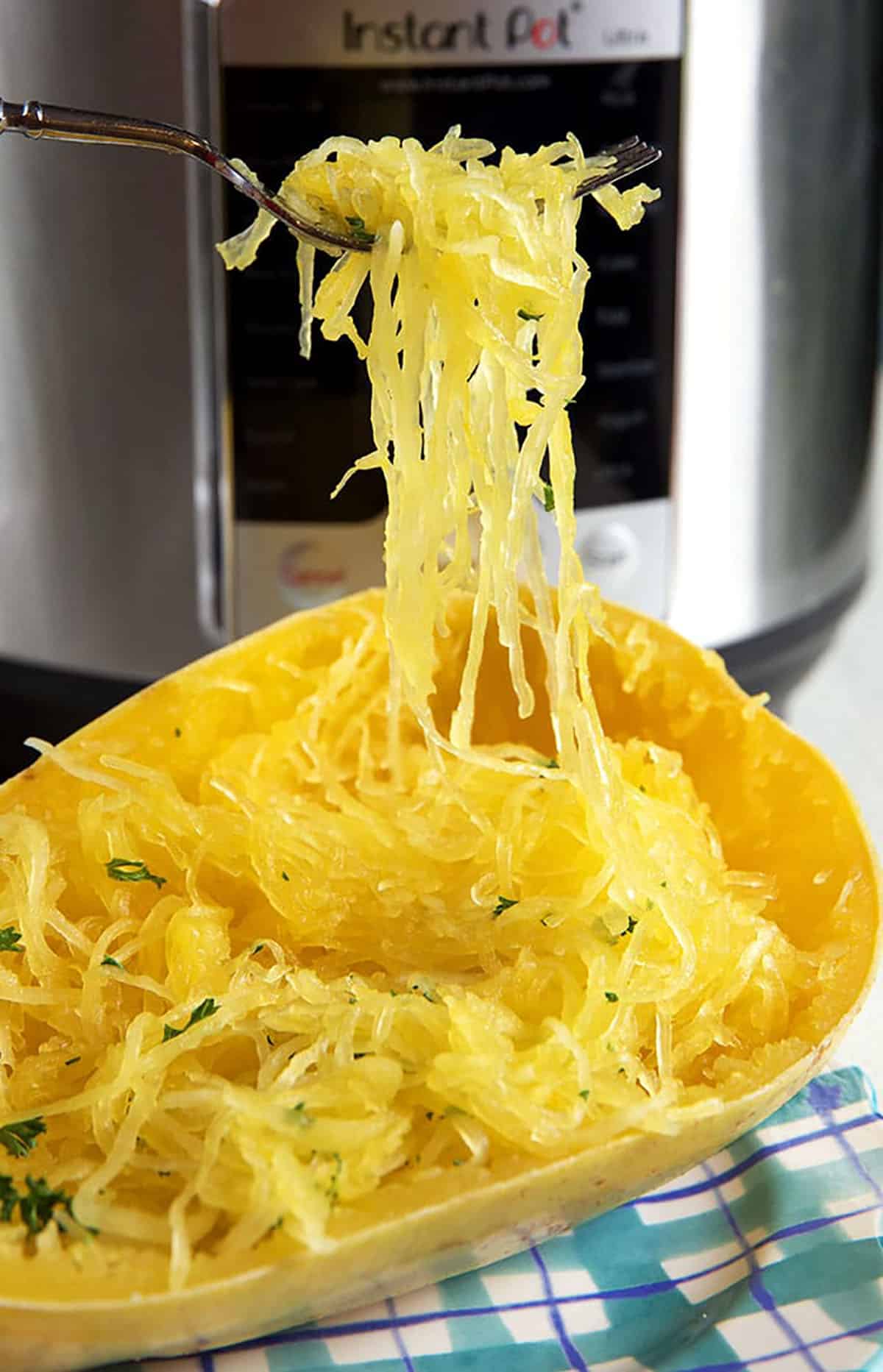 A fork is holding up a bite of spaghetti squash in front of an Instant Pot.