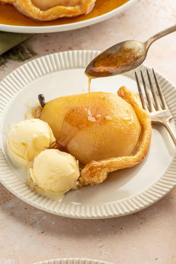 Slice of Pear Tarte Tatin on a white plate with two scoops of vanilla ice cream and a spoon drizzling the pan syrup over the pear.