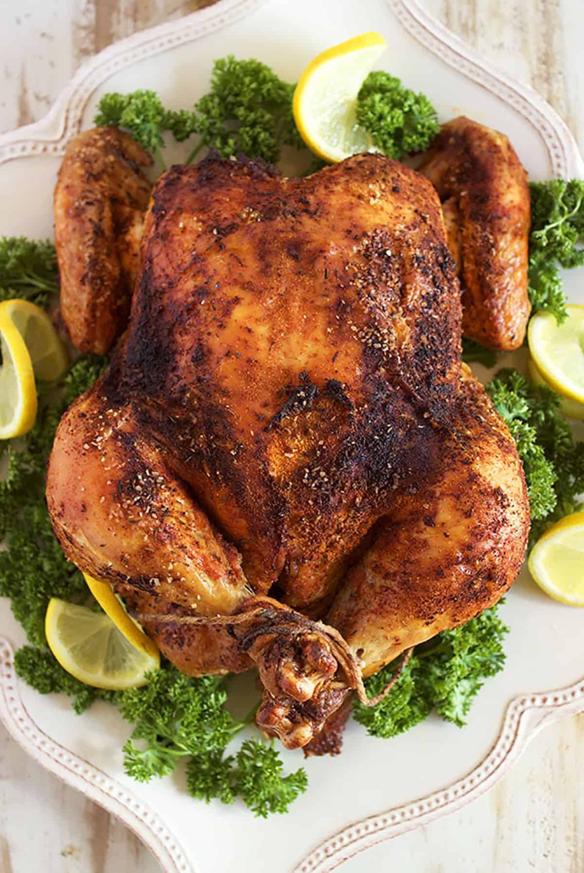 Whole roasted Rotisserie Chicken on a white platter with platter and slices of lemon.