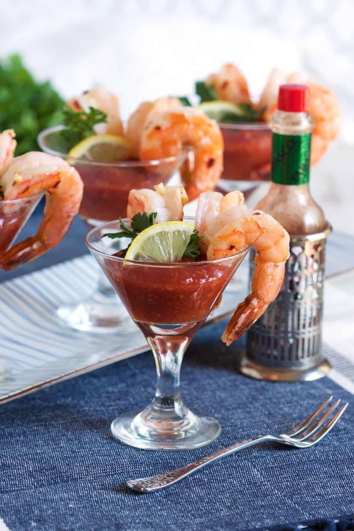 Shrimp cocktail plated on a mini martini glass filled with cocktail sauce and a bottle of tabasco in the background.