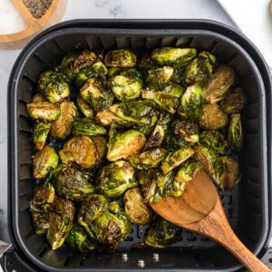 A wooden spoon is stirring come cooked brussels sprouts in an air fryer basket.