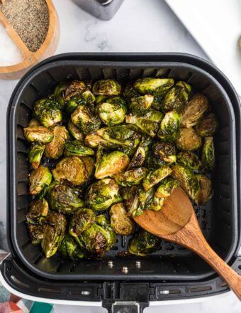 A wooden spoon is stirring come cooked brussels sprouts in an air fryer basket.